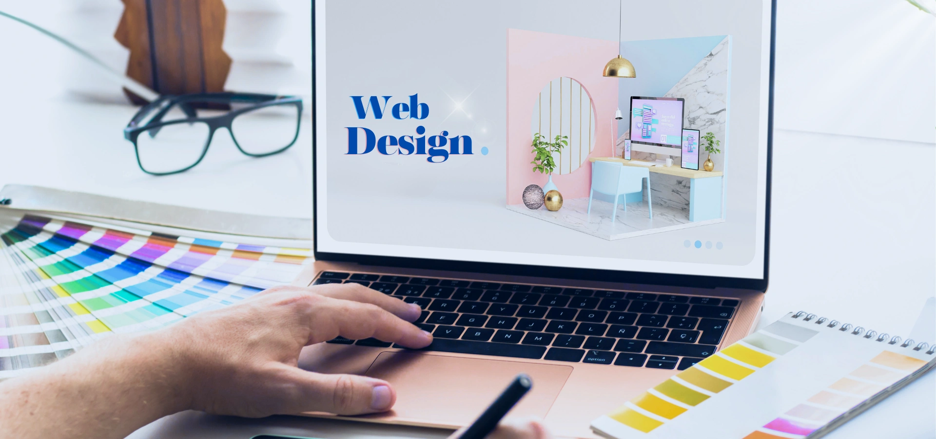 A person is using a rose-gold laptop to display a web design interface. The screen reads "Web Design", and a design palette can also be seen nearby.