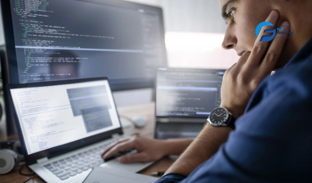 طرق الربح من التدوين A person sits at a desk with two laptops and a monitor, all displaying code. The person has their hand to their face, as though they are deep in thought.